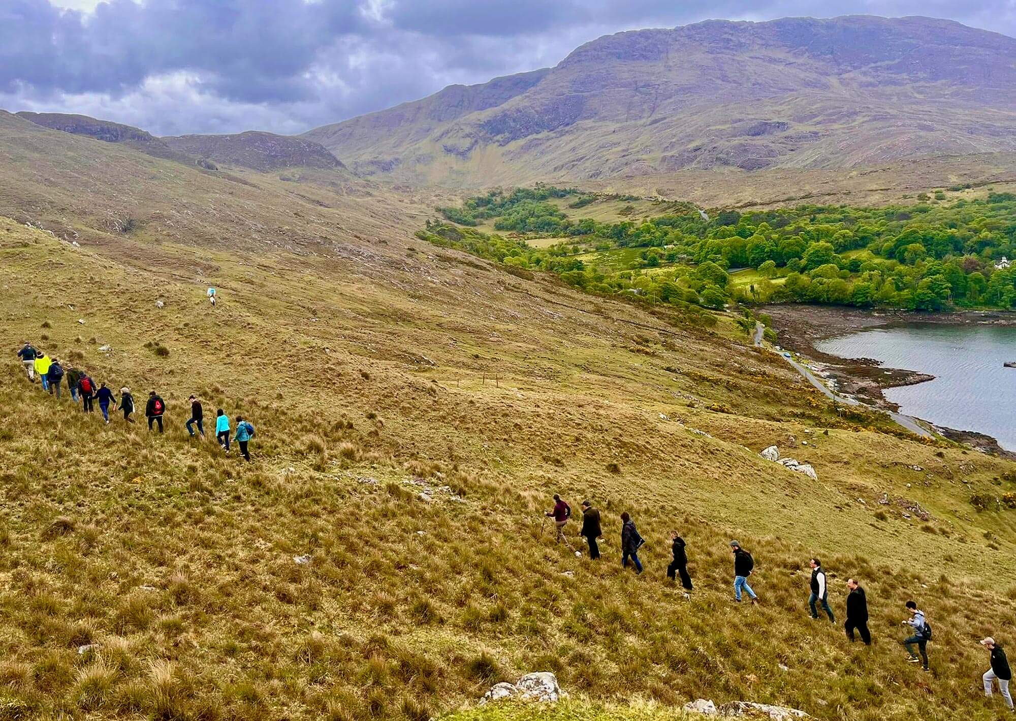Hikers in colorful jackets walk uphill on a golden-brown slope. Mountains and a lake are visible under an overcast sky.