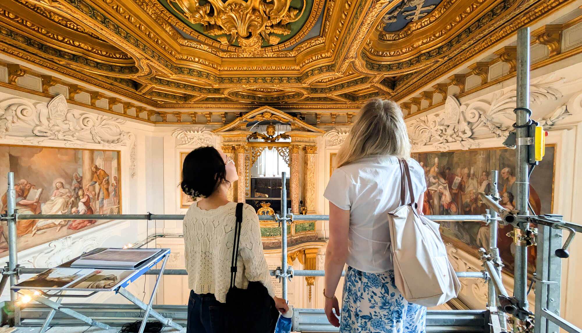 Two women on scaffolding gaze up at an opulent gold and blue gilded ceiling and wall paintings in a grand hall.