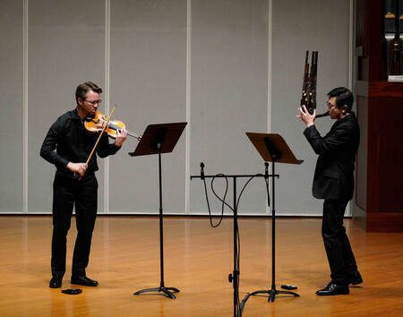 Two male musicians perform on stage. One plays a violin, the other plays a sheng. Both wear black clothing.
