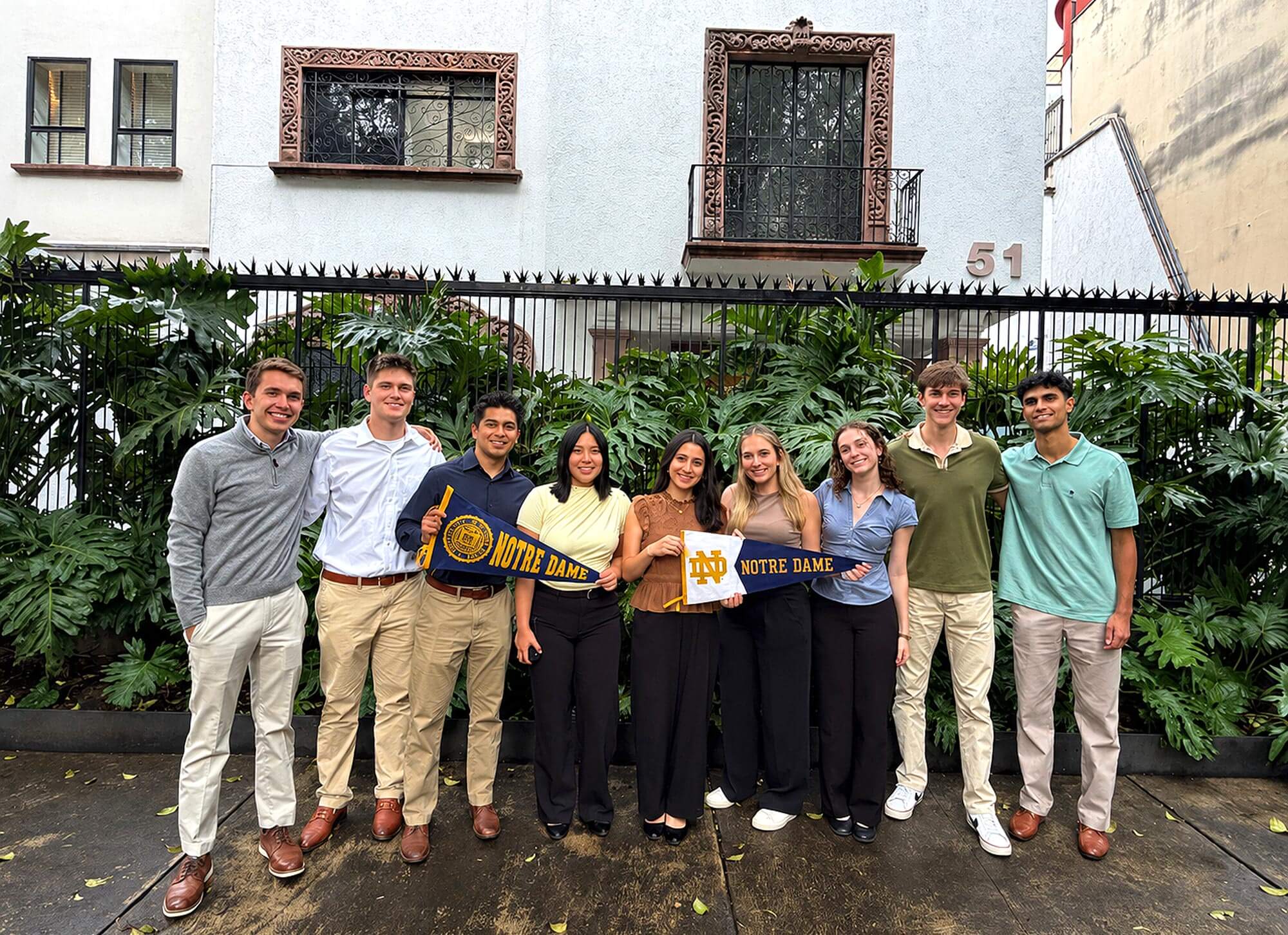 Nine smiling students pose outdoors, some holding Notre Dame pennants, in front of a white building with ornate windows.