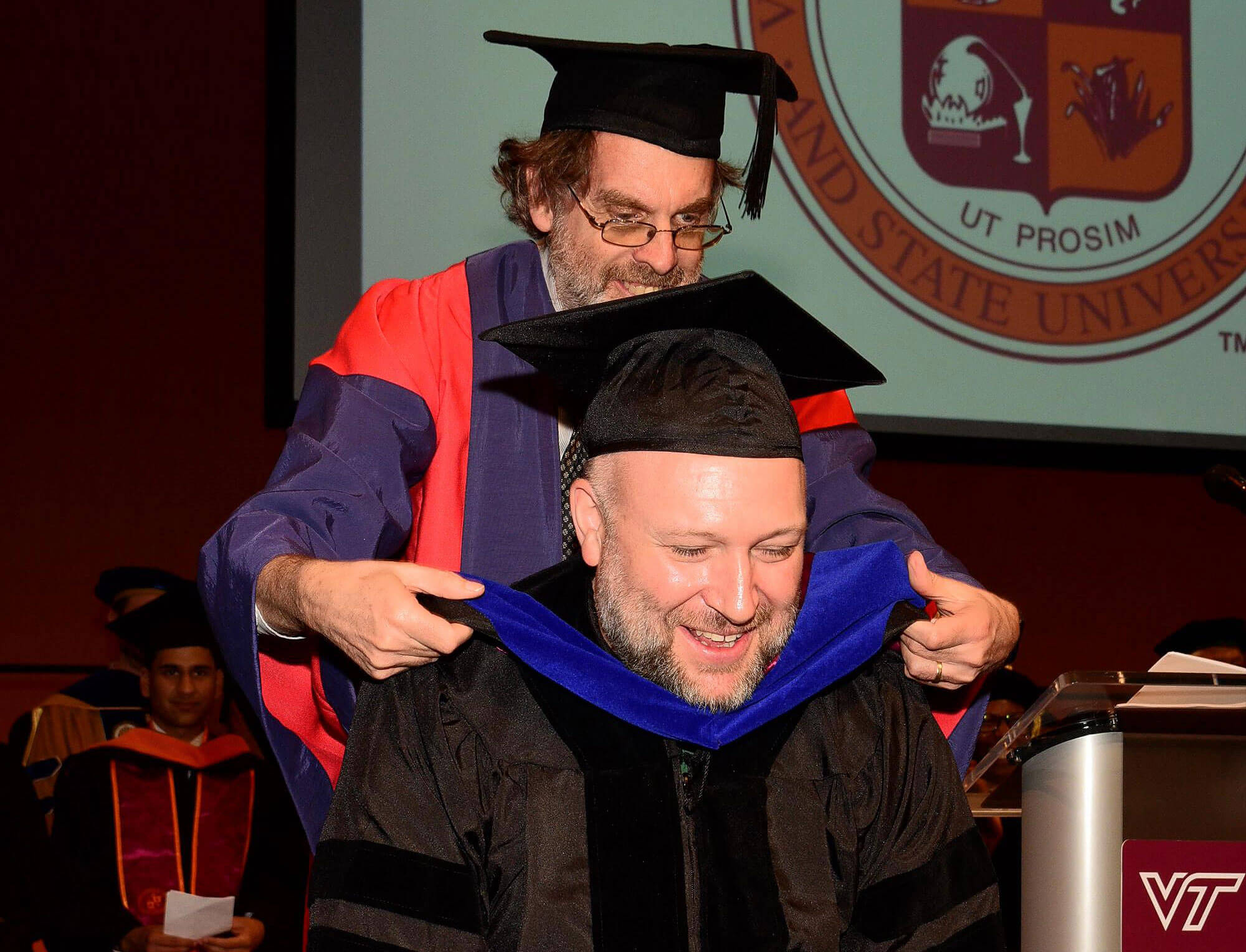 One man in academic robes places a blue velvet hood on a smiling man in black graduation attire.
