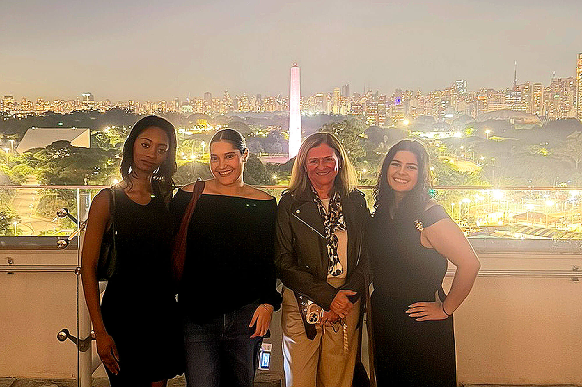 Four smiling women on a balcony at night, overlooking the São Paulo skyline with the illuminated Obelisco do Ibirapuera.