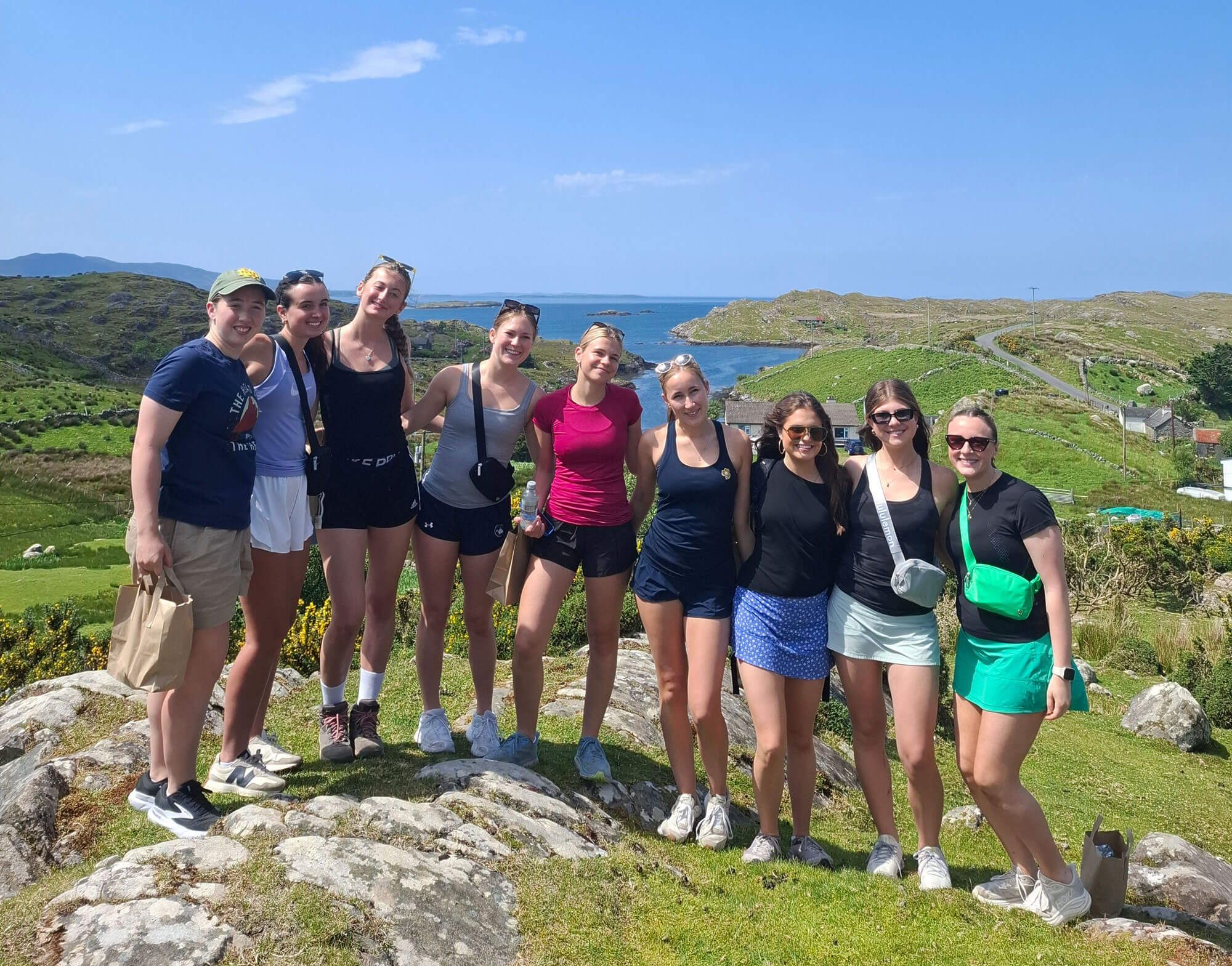 Eight smiling young women in athletic wear pose on a rocky, grassy hill overlooking a sunny blue ocean. One wears an ND logo.