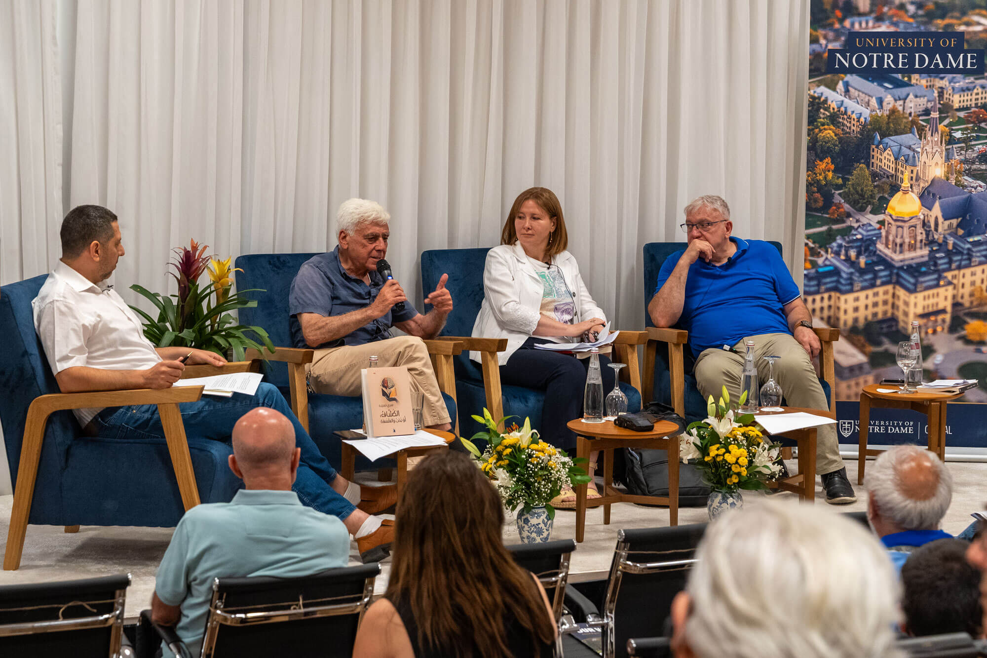 A white-haired man in a blue shirt speaks into a microphone on a panel with three others. A Notre Dame campus banner is visible.