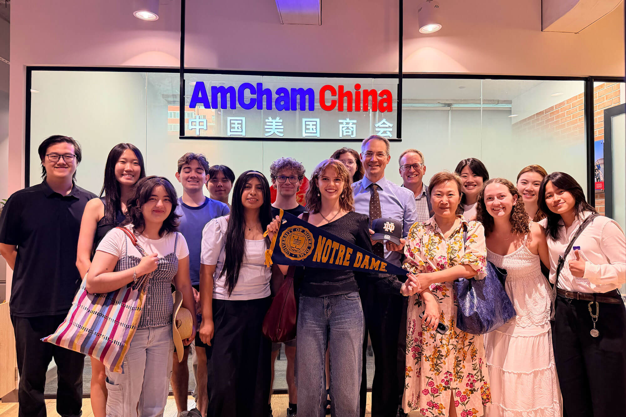 Smiling students and adults stand by the AmCham China sign. A woman holds a Notre Dame pennant, a man holds an ND hat.