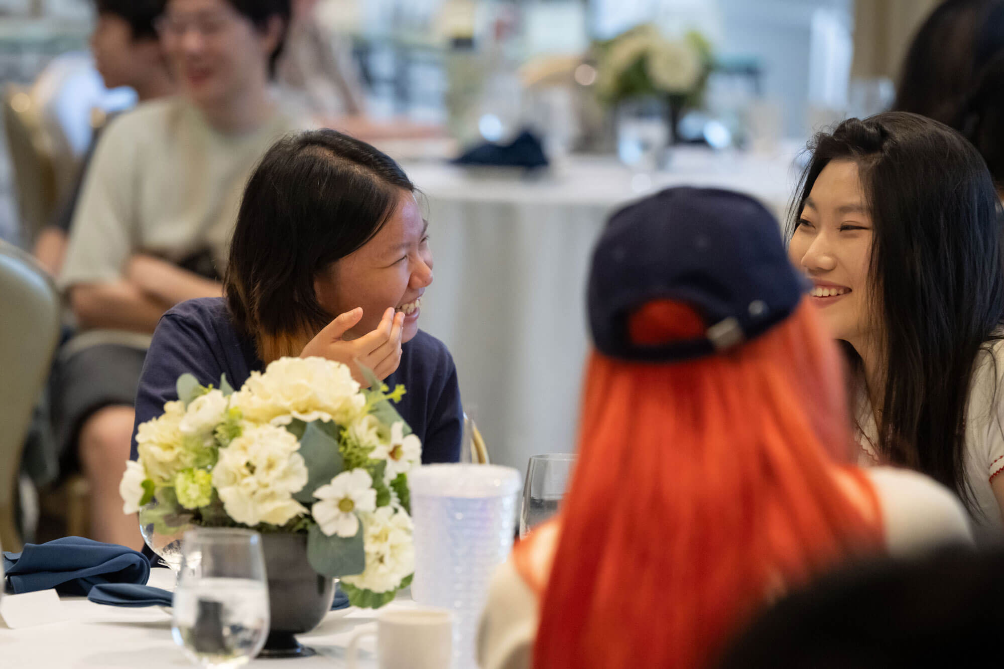 Two young women smile and laugh while conversing at a table with white floral centerpieces at a formal event.