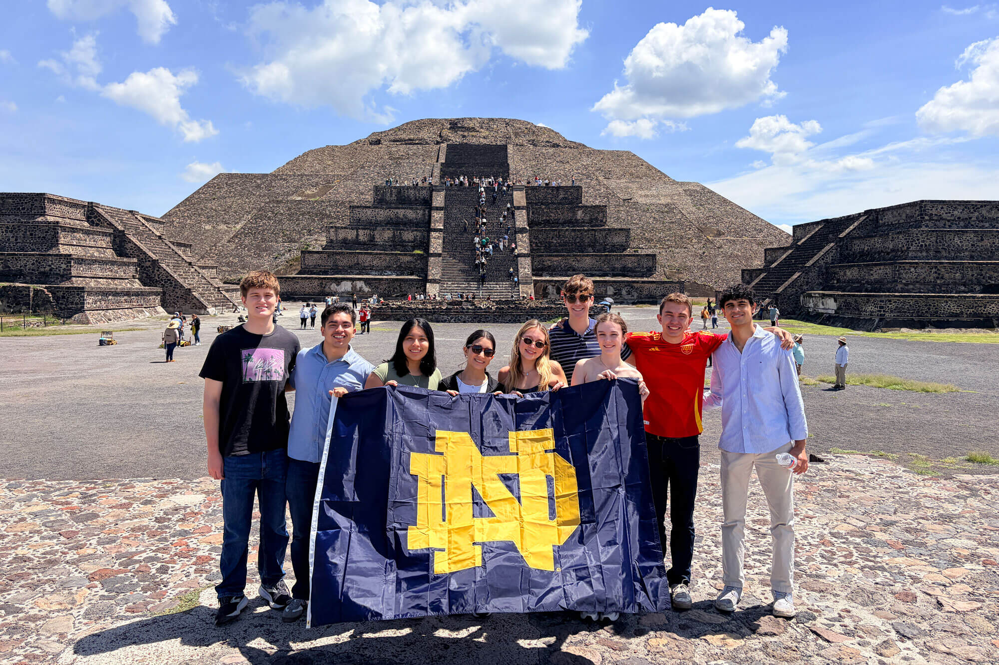Eight smiling students hold a navy blue and gold Notre Dame flag in front of the Pyramid of the Sun at Teotihuacan.