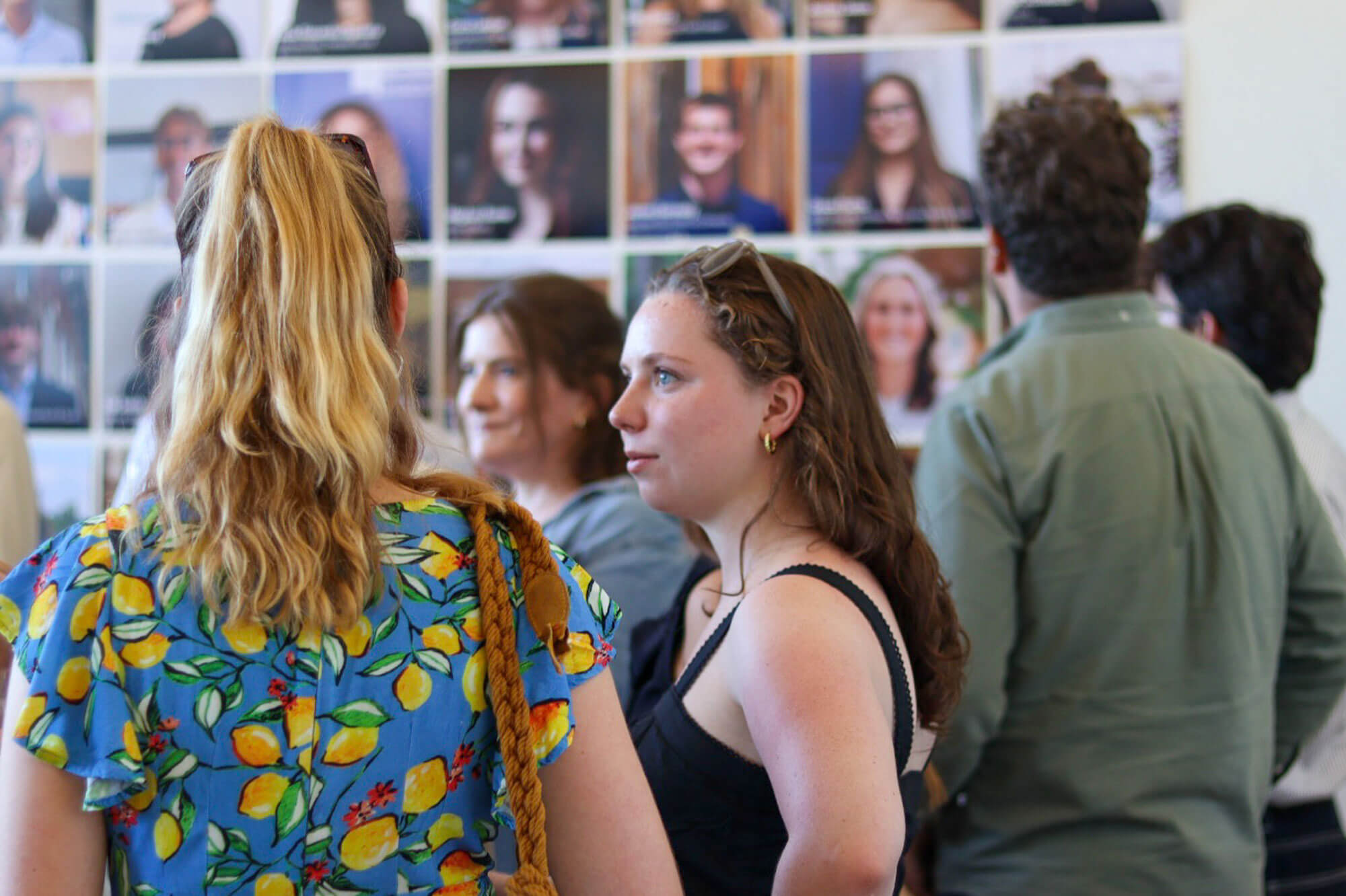Crowd views a wall of portraits. Prominent: woman in lemon dress, woman in black, man in green shirt facing wall.