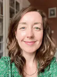 Smiling woman with wavy brown hair, one blue eye and one green eye, wearing a green patterned top.