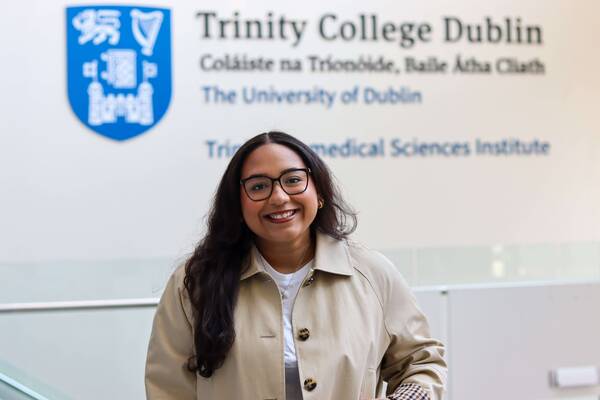 Smiling woman with dark hair and glasses in a tan coat, standing before the Trinity College Dublin wall logo and text.