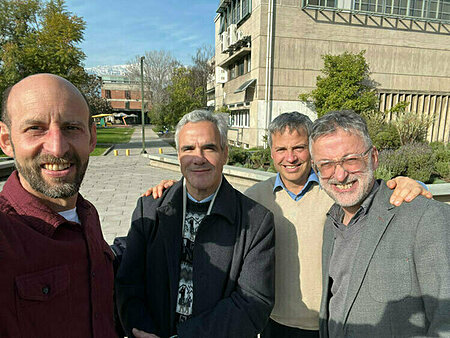 Four smiling men, varying ages and attire, pose for a selfie outdoors with modern buildings and distant mountains.