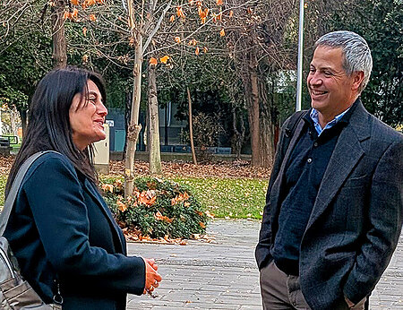 A dark-haired woman in a dark jacket smiles at a gray-haired man in a gray plaid blazer, laughing outdoors on a fall campus path.