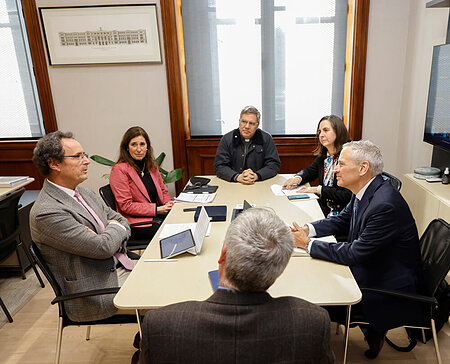 Six people, including a priest, discuss around a conference table. A man in a blue suit smiles, a woman wears a pink blazer.
