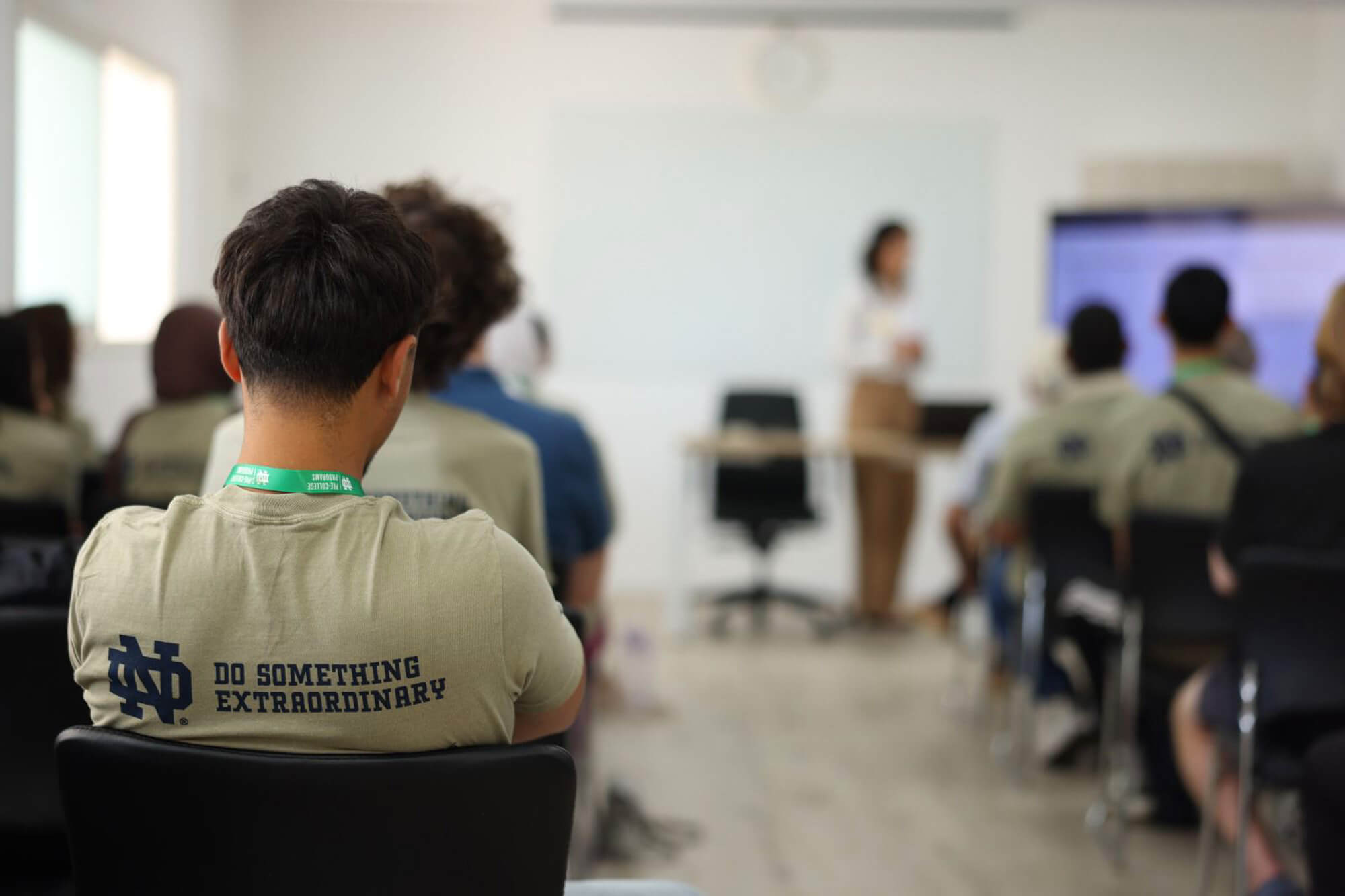 A person in a tan shirt with Notre Dame logo and "DO SOMETHING EXTRAORDINARY" faces away from the camera in a classroom.