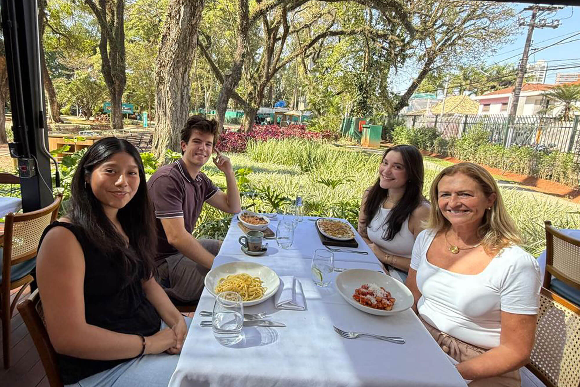 Four people smile at a table with pasta dishes and drinks outdoors.