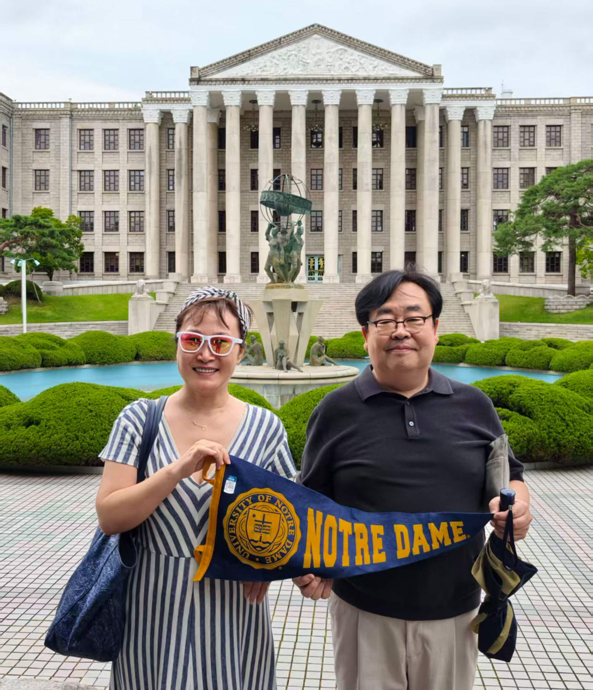 A couple smiles in front of a grand building, holding a Notre Dame pennant.