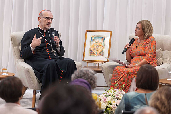 Cardinal wearing a cassock and cross speaks to a woman in an orange dress holding a microphone.