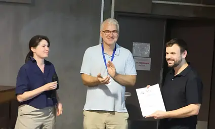 Two men and a woman smile as one man accepts a certificate and another claps.